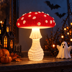 A glowing mushroom-shaped table lamp with a red cap and white spots sits on a wooden surface in a Halloween-themed room setting with pumpkins, a ghost figurine, and warm string lights in the background.