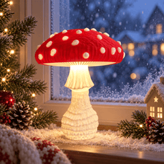 A mushroom-shaped table lamp with a red cap and white spots, glowing warmly on a snowy windowsill decorated with pine branches, pinecones, and a small house ornament.