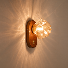 A wall-mounted light fixture with a warm wood backplate and a faceted clear glass shade glowing with a warm amber light, mounted on a beige wall with light rays casting outward.