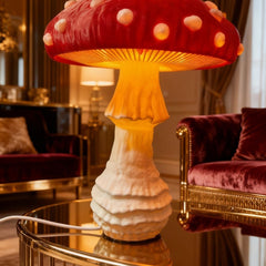 A mushroom-shaped table lamp with a glossy red cap, white spots, and a glowing warm yellow-orange stem and underside, placed on a reflective gold table in an elegant living room with velvet furniture.
