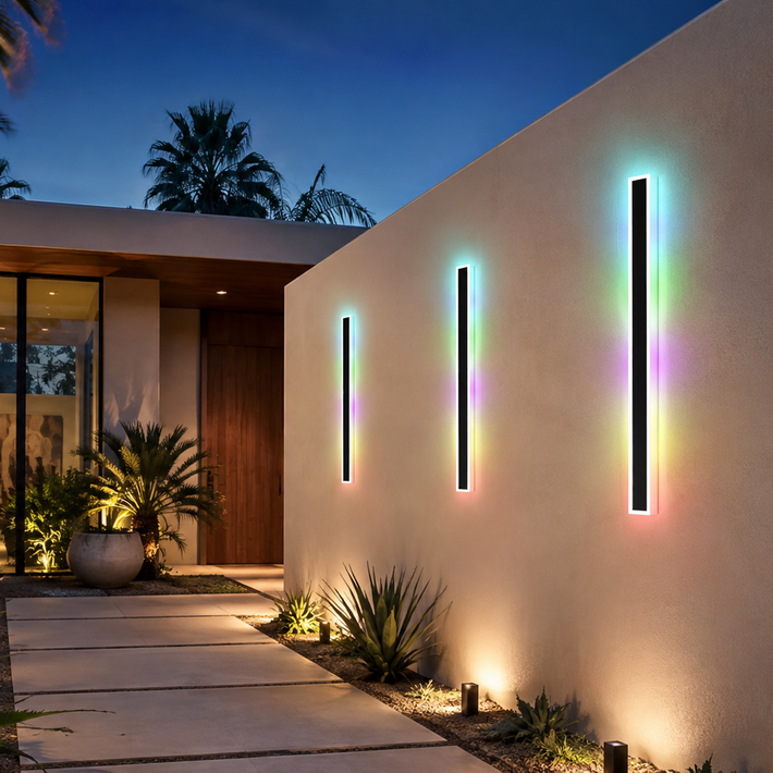 Three vertical black outdoor wall light fixtures with rainbow LED lighting mounted on a beige stucco wall of a modern house at dusk.
